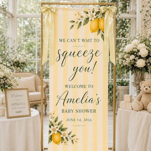Puede incluir: Un cartel rectangular alto con rayas verticales amarillas y blancas, con ilustraciones de limones y flores. El texto dice: WE CAN'T WAIT TO squeeze you! WELCOME TO Amelia's BABY SHOWER JUNE 14, 2026. Sujeto a un marco de metal dorado con lazos.