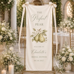 May include: A vertical cream-colored fabric banner hanging from a gold metal stand with satin ribbons. The banner features a watercolor illustration of two pears with leaves and text reading: THE Perfect PEAR, WELCOME TO Charlotte's BRIDAL SHOWER 04.18.2027.