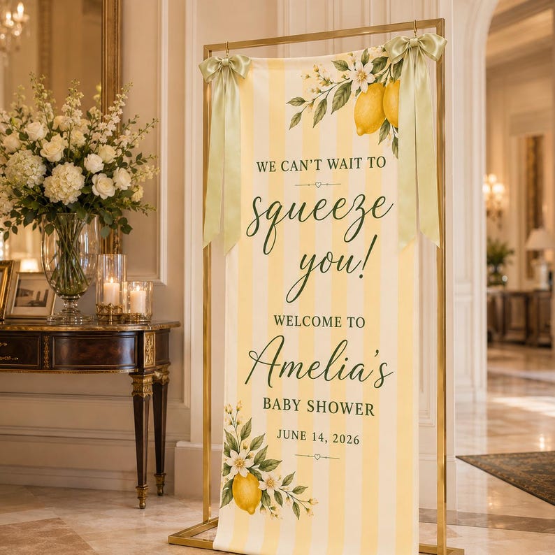 Puede incluir: Un cartel rectangular vertical con rayas amarillas y blancas, decorado con ilustraciones de limones y flores blancas. El texto dice: WE CAN'T WAIT TO squeeze you! WELCOME TO Amelia's BABY SHOWER JUNE 14, 2026. Est&aacute; colgado de un marco de metal dorado.