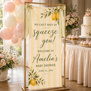 Puede incluir: Un cartel rectangular vertical con rayas amarillas y blancas, decorado con ilustraciones de limones y flores blancas. El texto dice: WE CAN'T WAIT TO squeeze you! WELCOME TO Amelia's BABY SHOWER JUNE 14, 2026. Est&aacute; colgado de un marco de metal dorado.