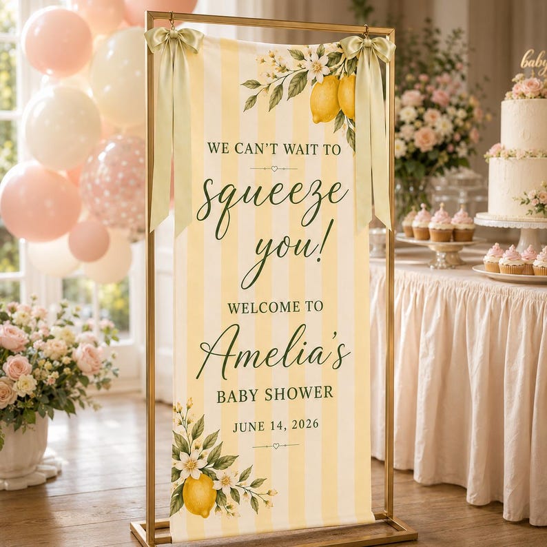 Puede incluir: Un cartel rectangular vertical con rayas amarillas y blancas, decorado con ilustraciones de limones y flores blancas. El texto dice: WE CAN'T WAIT TO squeeze you! WELCOME TO Amelia's BABY SHOWER JUNE 14, 2026. Est&aacute; colgado de un marco de metal dorado.