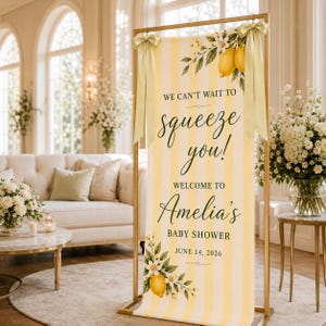 Puede incluir: Un cartel vertical con rayas amarillas y blancas, decorado con ilustraciones de limones y flores. El texto dice: WE CAN'T WAIT TO squeeze you! WELCOME TO Amelia's BABY SHOWER JUNE 14, 2026. El cartel est&aacute; sujeto por un marco de color dorado.