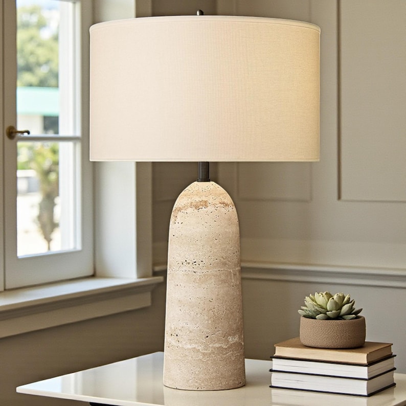 May include: A beige table lamp with a cylindrical stone base and a cream-colored lampshade. The lamp is on a white table next to a small succulent in a brown pot and a stack of books. The lamp is lit.