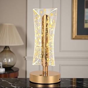 May include: A gold-colored table lamp with a clear, curved shade adorned with gold leaf accents. The lamp has a cylindrical gold base and a central gold column. The lamp is designed for indoor use and adds a touch of elegance to any room.