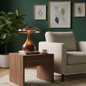 May include: A modern wooden table lamp with a brown base and a clear, rounded top. The lamp sits on a wooden side table next to a cream-colored armchair. A small teacup and books are also on the table. The background features framed artwork and a green wall.