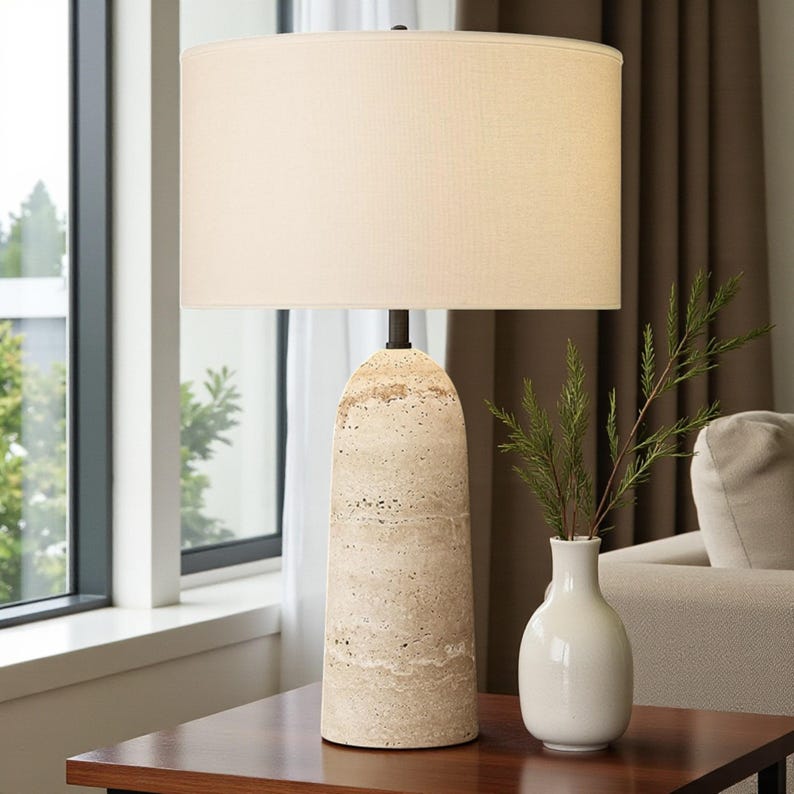 May include: A beige table lamp with a cylindrical stone base and a cream-colored lampshade. The lamp is on a dark wooden table. A white vase with green sprigs sits next to the lamp. The lamp is approximately 28 inches tall.