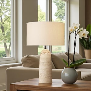 May include: A cream-coloured table lamp with a cylindrical, textured base and a lampshade. The lamp is on a dark wood table, next to a small grey vase with white orchids. The background includes a beige sofa and a window.