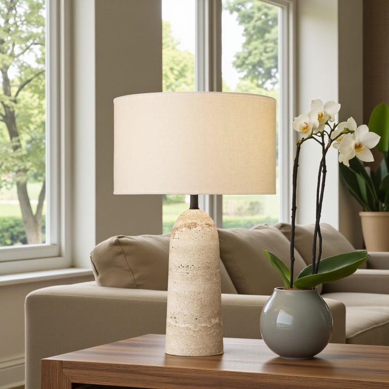 May include: A beige table lamp with a cylindrical, textured base and a cream-colored lampshade. The lamp sits on a dark wood table, next to a small gray vase with white orchids. The background includes a beige sofa and a window.