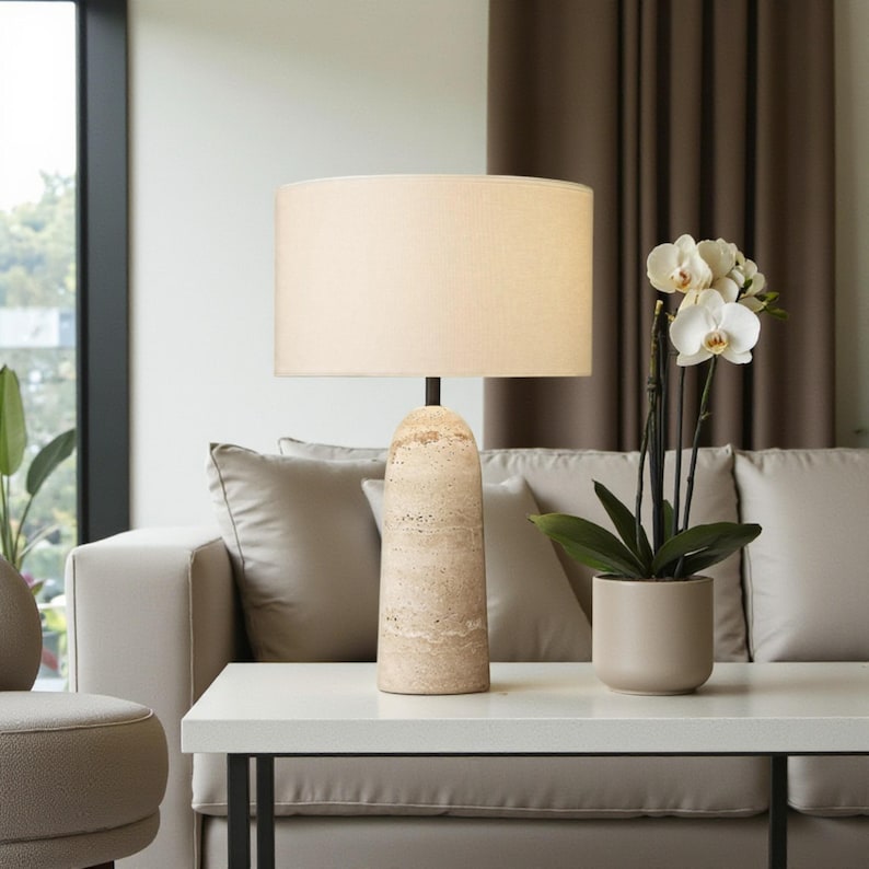 May include: A beige table lamp with a cylindrical stone base and a cream-colored lampshade. The lamp is on a white table, with a beige sofa and a potted orchid in the background. The lamp's height is approximately 24 inches.