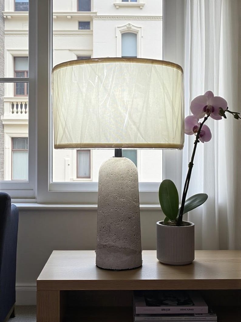May include: A modern table lamp with a textured, light gray concrete base and a cream-colored lampshade. A potted orchid with pink flowers sits nearby. The lamp is on a wooden table near a window.