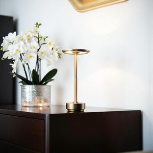 May include: A gold table lamp with a circular shade and base, and a slender stem. The lamp is on a dark brown surface, next to a silver pot with white flowers. A gold-framed mirror hangs on the white wall.