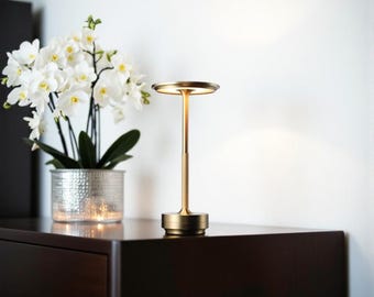 May include: A gold-colored table lamp with a circular shade and base, and a slender stem. The lamp is on a dark brown surface, next to a silver pot with white flowers. A gold-framed mirror hangs on the white wall.