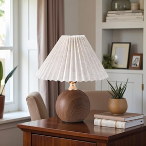 May include: A table lamp featuring a pleated cream lampshade. The lamp has a spherical, wooden base with a gold-coloured accent. It sits on a wooden table, next to a small potted plant and books.