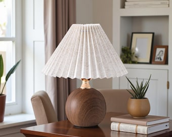 May include: A decorative table lamp with a pleated off-white lampshade. The lamp has a round, wooden base with a gold-colored accent. The lamp is on a wooden table, next to a small potted plant and books.