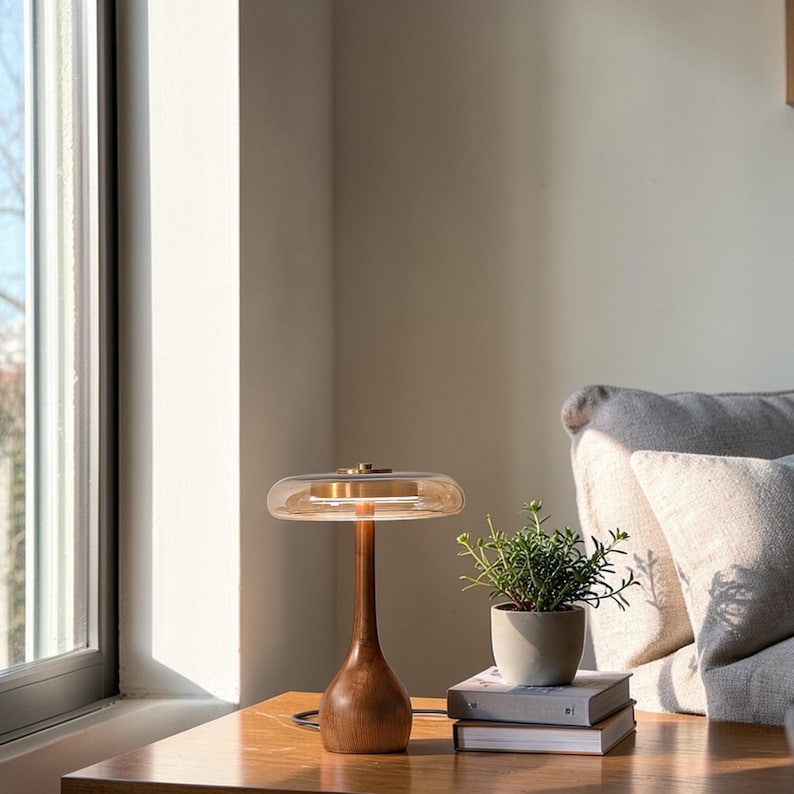 May include: A wooden table lamp with a brown base and a clear, rounded shade. The lamp has a brass accent and sits on a wooden table. A small potted plant and books are next to the lamp. The room has a window and a gray couch.