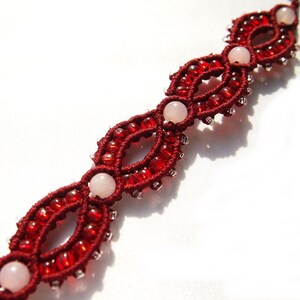 May include: A handmade macrame bracelet featuring a deep red color scheme. The bracelet is adorned with small red beads and larger, pale pink, round gemstones. The intricate design showcases a series of connected oval shapes.