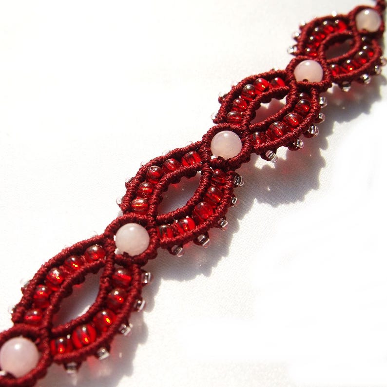 May include: A handmade macrame bracelet featuring a deep red color scheme. The bracelet is adorned with small red beads and larger, pale pink, round gemstones. The intricate design showcases a series of connected oval shapes.