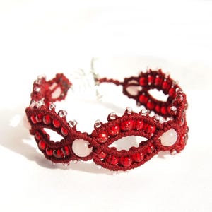May include: A handmade macrame bracelet featuring a deep red cord and accented with red and clear beads. The bracelet also includes three light pink, round beads. The intricate design is visible against a white background.