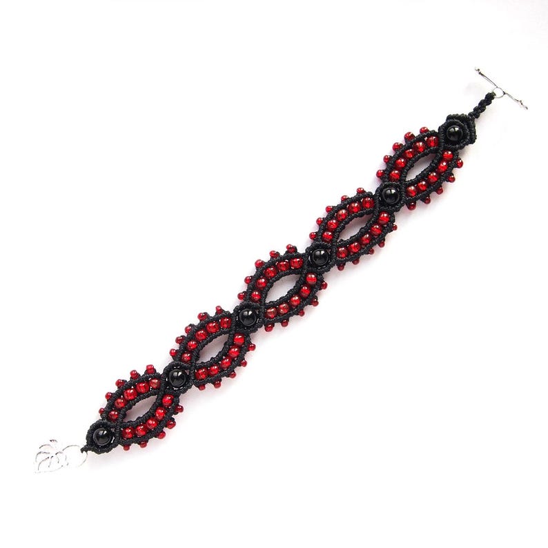 May include: A macrame bracelet featuring a repeating pattern of red beads and black knots. The bracelet has a toggle clasp and a silver-colored charm at one end. The design incorporates black beads at intervals, creating a striking contrast.