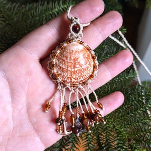 Puede incluir: Un collar con colgante de concha hecho a mano. El colgante presenta una concha marr&oacute;n y blanca, intrincadamente tejida con cord&oacute;n beige y adornada con cuentas de color &aacute;mbar. Peque&ntilde;os dijes de ancla dorados cuelgan de la parte inferior.