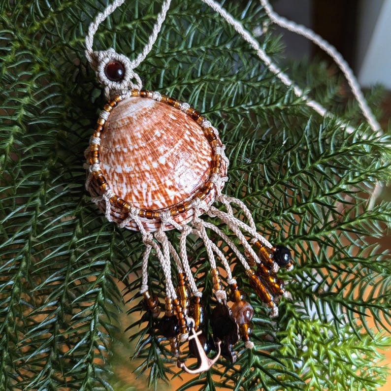 Puede incluir: Un collar de macram&eacute; hecho a mano con un gran colgante de concha. La concha es marr&oacute;n y blanca, rodeada por un borde de cuentas. El collar tiene un cord&oacute;n color crema, cuentas marrones y un peque&ntilde;o dije de ancla.