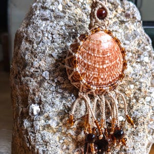 Puede incluir: Un colgante artesanal de concha con un dise&ntilde;o a rayas marr&oacute;n y blanco. La concha est&aacute; encerrada en una red de macram&eacute;, adornada con cuentas de color &aacute;mbar y detalles colgantes. El colgante cuelga de un cord&oacute;n beige.