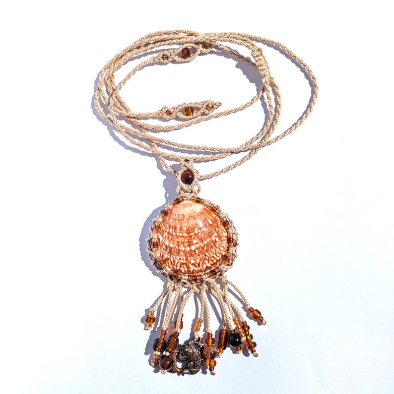 Puede incluir: Collar de macram&eacute; hecho a mano con un colgante de concha. El colgante es una concha grande y estampada, rodeada de cuentas marrones. Cuelga de un cord&oacute;n beige. Cuentas marrones y un peque&ntilde;o dije cuelgan debajo.