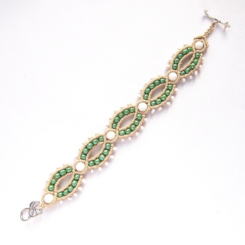 May include: Macrame bracelet featuring a repeating pattern of green and white beads. The bracelet is made with beige cord and has a silver-tone clasp. The design includes oval-shaped sections of green beads separated by white beads.
