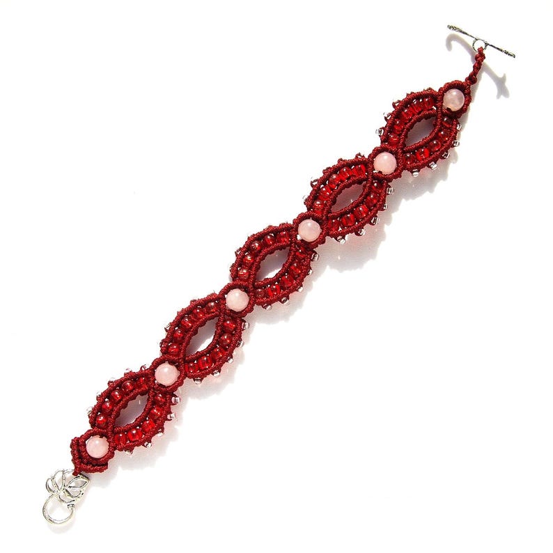 May include: A handmade macrame bracelet in a deep red color, featuring an intricate woven design. The bracelet is adorned with small, clear beads and light pink gemstone accents. It has a silver-tone clasp and is displayed on a white background.