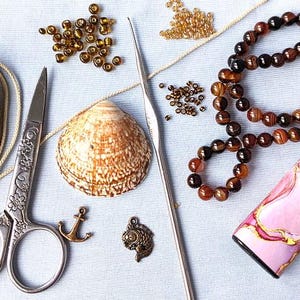 Puede incluir: Una colecci&oacute;n de materiales de artesan&iacute;a que incluye una concha, tijeras, cuentas, un ganchillo, una pulsera y un estuche decorativo. Los art&iacute;culos est&aacute;n dispuestos sobre una superficie azul claro, lo que sugiere un tema de artesan&iacute;a o fabricaci&oacute;n de joyas.