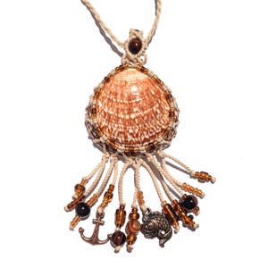 Puede incluir: Un collar con colgante de concha hecho a mano. El colgante presenta una concha marr&oacute;n y blanca, encerrada en una red de macram&eacute;, adornada con cuentas y abalorios marrones, incluyendo un ancla y un pez. El collar est&aacute; en un cord&oacute;n beige.