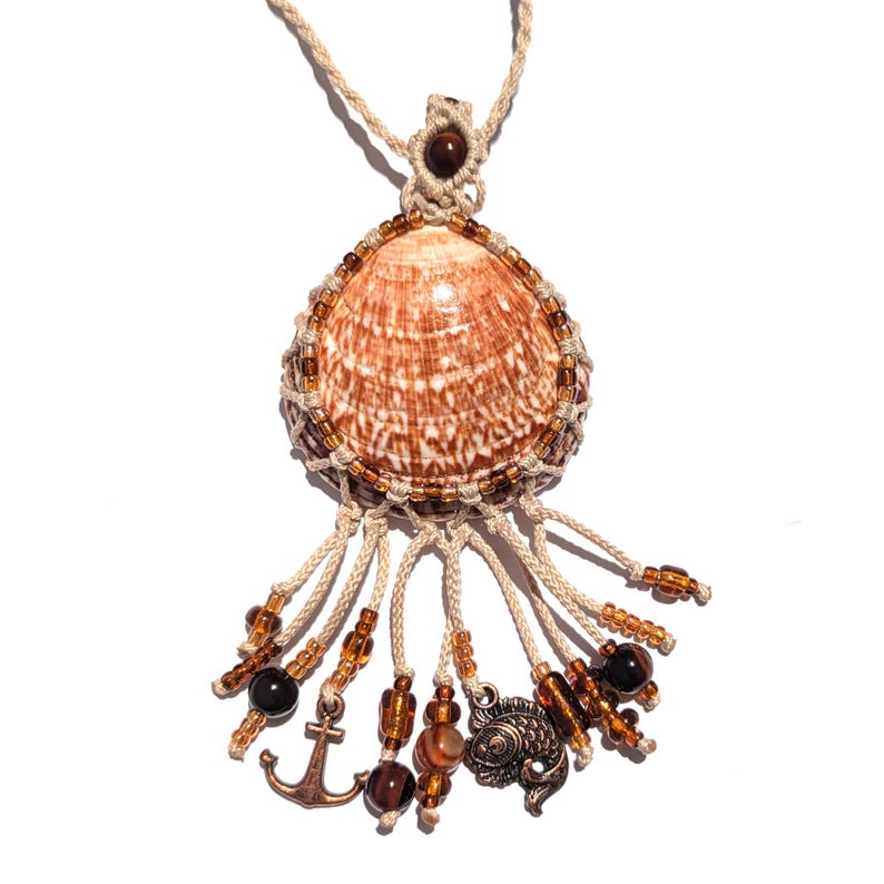 Puede incluir: Un collar con colgante de concha hecho a mano. El colgante presenta una concha marr&oacute;n y blanca, encerrada en una red de macram&eacute;, adornada con cuentas y abalorios marrones, incluyendo un ancla y un pez. El collar est&aacute; en un cord&oacute;n beige.