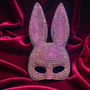 May include: A pink bunny mask covered in iridescent rhinestones. The mask has large, upright ears and eye openings. The mask is set against a deep red velvet background.