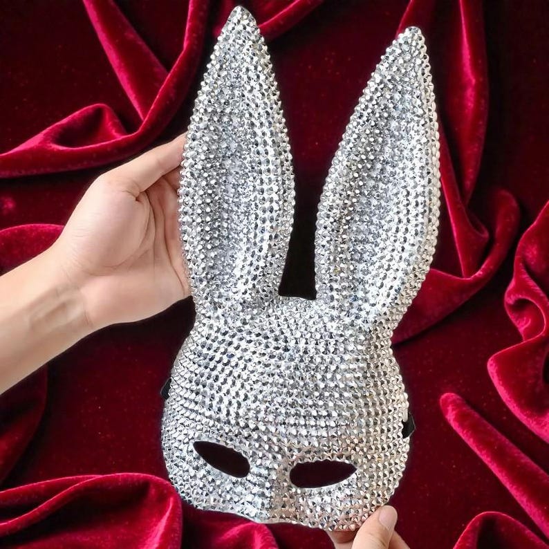 May include: A silver bunny mask covered in sparkling rhinestones. The mask has long, upright ears and eye openings. The mask is held against a deep red velvet background.