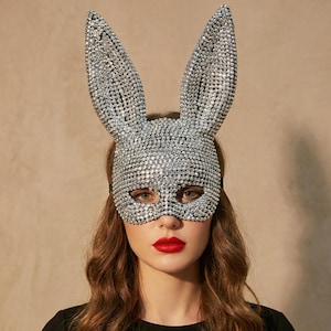 May include: A silver bunny mask with sparkling rhinestones. The mask features long, upright ears and covers the eyes and nose. The mask is worn by a person with red lipstick and brown hair.