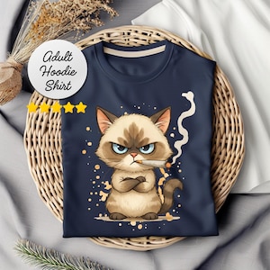 Puede incluir: Camiseta con capucha para adultos de color azul marino con un gato de dibujos animados gruñón con un cigarrillo. El gato tiene los brazos cruzados y una expresión enfadada. La camiseta se muestra en una cesta de mimbre.