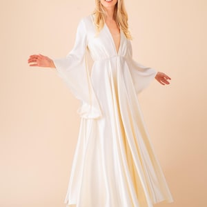 May include: A floor-length, ivory-colored dress with a plunging neckline and flowing bell sleeves. The dress has a fitted waist and a flared skirt. The model is wearing beige heels. The background is a solid peach color.