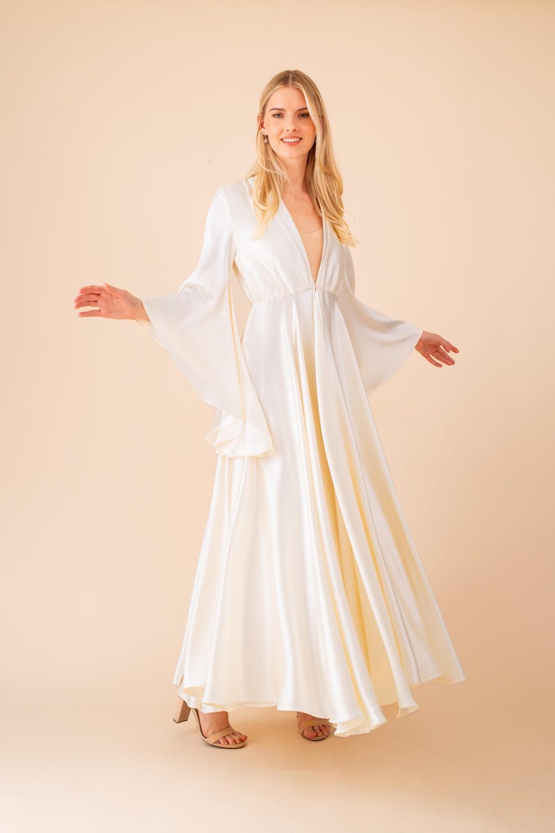 May include: A floor-length, ivory-colored dress with a plunging neckline and flowing bell sleeves. The dress has a fitted waist and a flared skirt. The model is wearing beige heels. The background is a solid peach color.