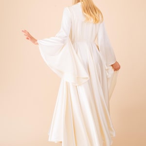 May include: A long, flowing ivory dress with wide, bell sleeves. The dress features a fitted bodice and a full skirt, creating an elegant silhouette. The model is viewed from the back, with blonde hair cascading down. The dress is paired with beige heels.
