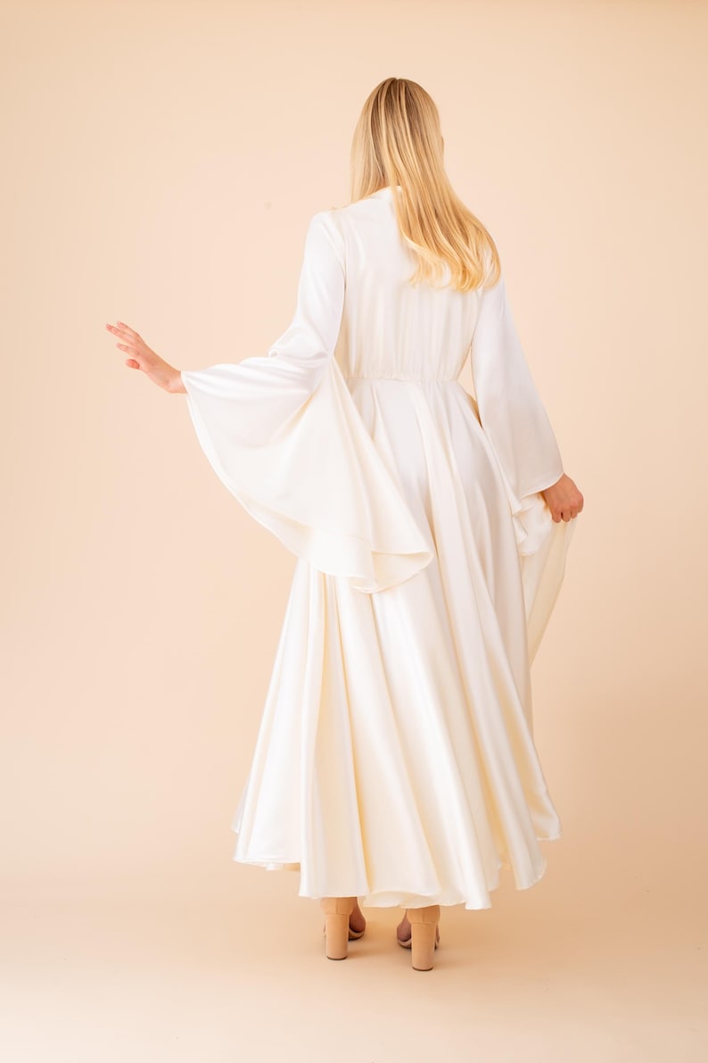 May include: A long, flowing ivory dress with wide, bell sleeves. The dress features a fitted bodice and a full skirt, creating an elegant silhouette. The model is viewed from the back, with blonde hair cascading down. The dress is paired with beige heels.