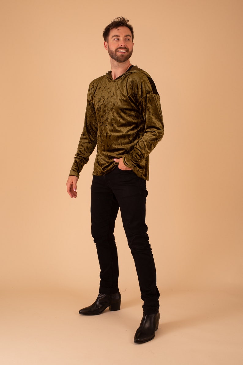 Op de afbeelding: Een man draagt een groen fluwelen hoodie met zwarte accenten, een zwarte jeans en zwarte laarzen met hak.
