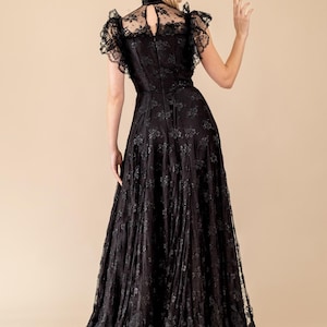 Pode incluir: Um vestido longo de renda preta com gola alta e mangas curtas com babados. O vestido apresenta intrincados padr&otilde;es florais e uma saia fluida. A modelo &eacute; vista por tr&aacute;s, com cabelo loiro, em um fundo neutro.