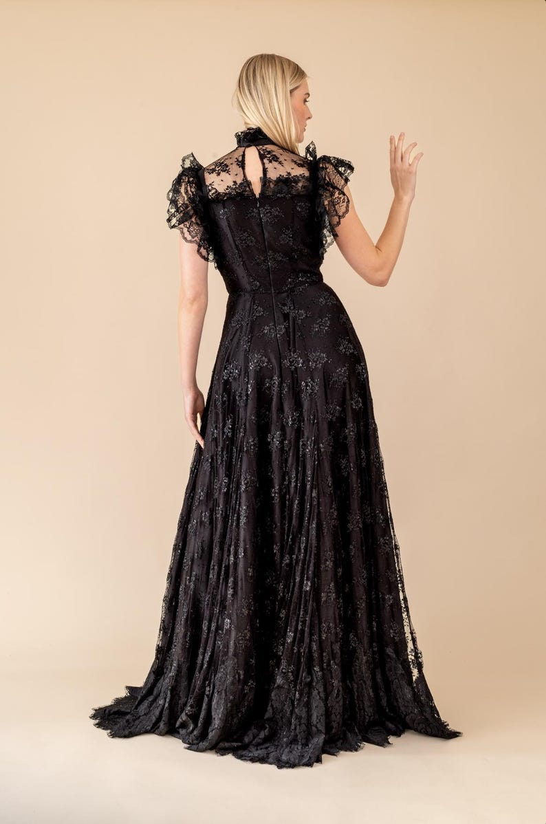 Pode incluir: Um vestido longo de renda preta com gola alta e mangas curtas com babados. O vestido apresenta intrincados padr&otilde;es florais e uma saia fluida. A modelo &eacute; vista por tr&aacute;s, com cabelo loiro, em um fundo neutro.