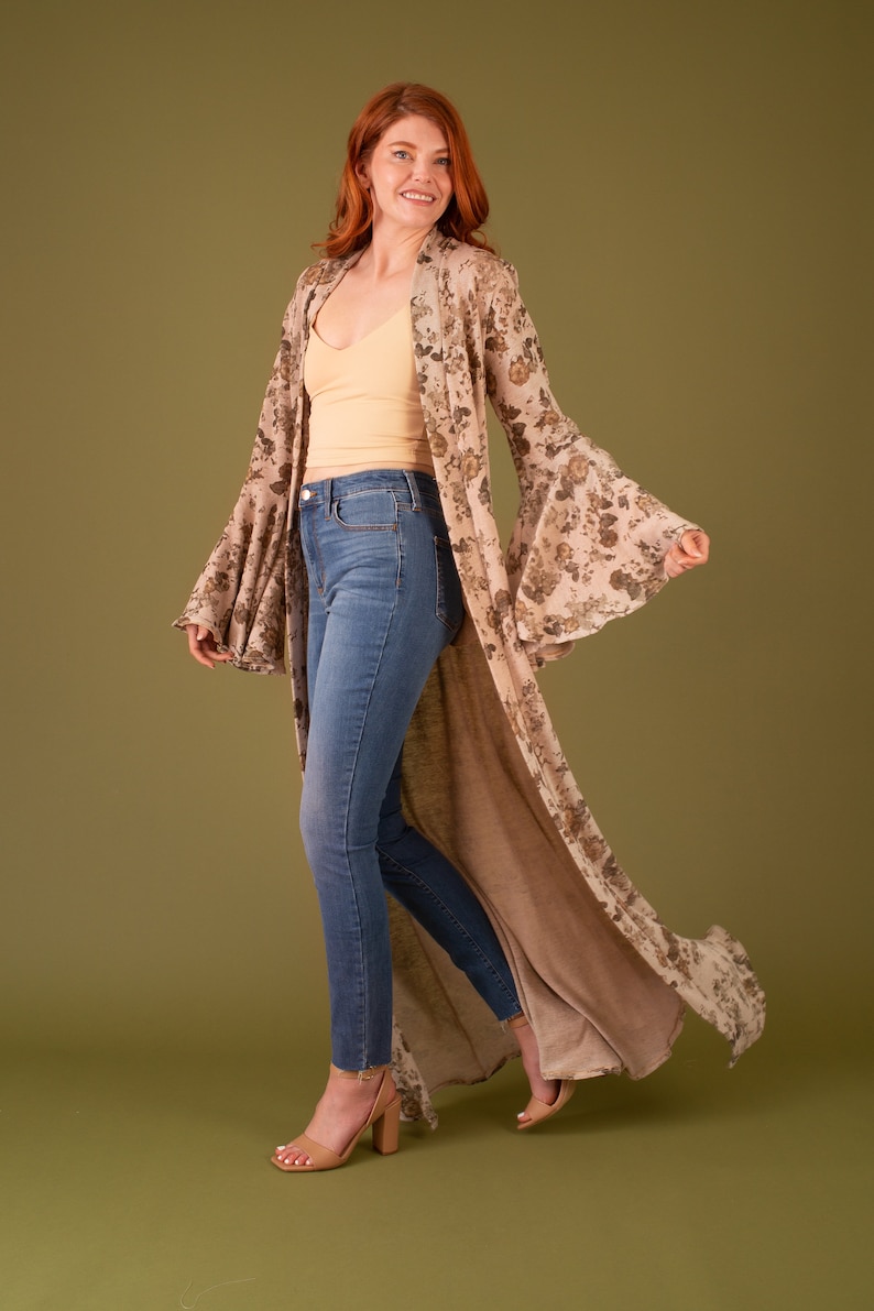 Puede incluir: Una mujer lleva una larga chaqueta de punto con estampado floral y mangas acampanadas sobre un top beige sin mangas y unos vaqueros azules. Lleva sandalias de tac&oacute;n nude.