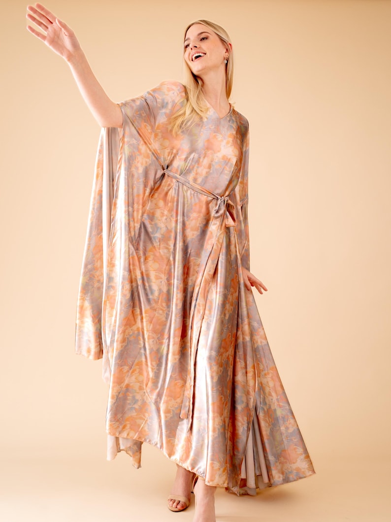May include: A long, flowing, peach-colored dress with a tie waist and a floral print. The dress is made of a silky material and has a relaxed fit.