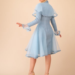 Op de afbeelding: Een lichtblauwe jurk met een cape en een tule overlay. De jurk heeft een getailleerd bovenstuk en een uitlopende rok. Het model draagt ​​nude hakken.
