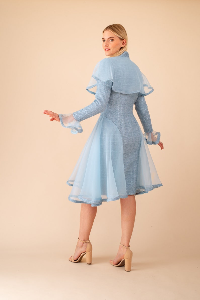 Op de afbeelding: Een lichtblauwe jurk met een cape en een tule overlay. De jurk heeft een getailleerd bovenstuk en een uitlopende rok. Het model draagt ​​nude hakken.