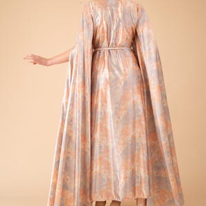 May include: A long, flowing dress with a cape-like design. The dress is made of a soft, silky fabric with a subtle shimmer and a light pink and gray floral pattern. The dress has a cinched waist and a flowing skirt.