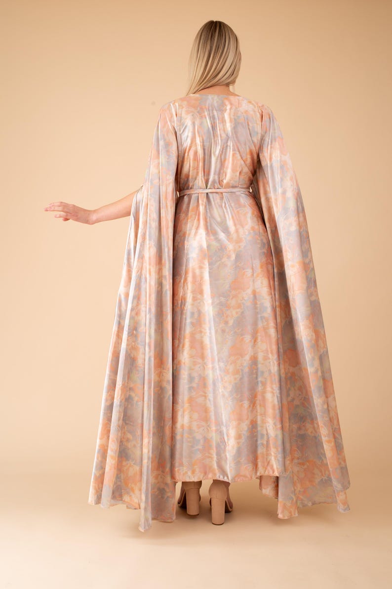May include: A long, flowing dress with a cape-like design. The dress is made of a soft, silky fabric with a subtle shimmer and a light pink and gray floral pattern. The dress has a cinched waist and a flowing skirt.
