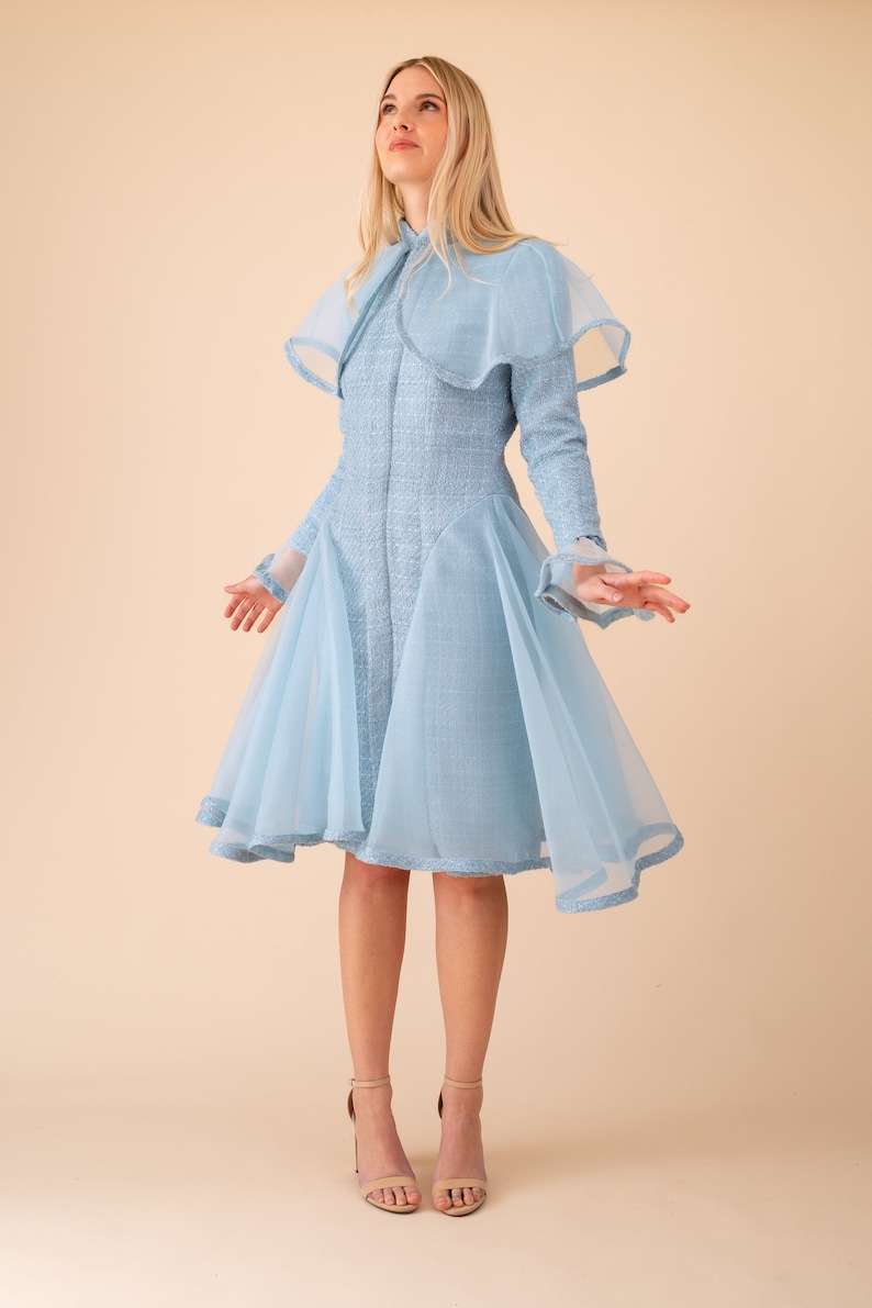 Op de afbeelding: Een lichtblauwe jurk met een transparante overlay en een hoge halslijn. De jurk heeft een getailleerd bovenlijf en een zwierige rok. Het model draagt de jurk met nude hakken.